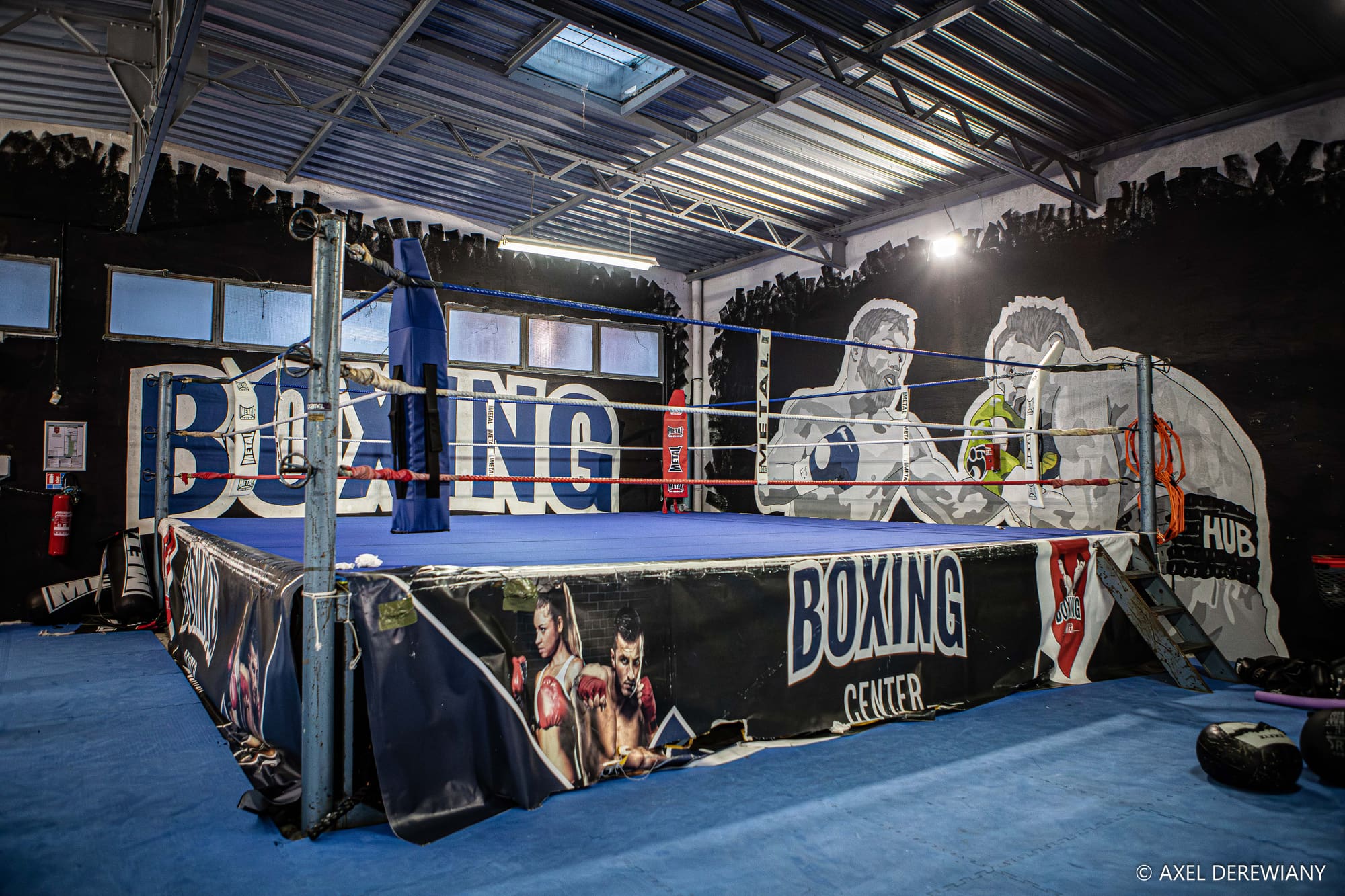 Le ring au tablier « Boxing Center », cordes rouges et bleues, devant la fresque murale de deux boxeurs peinte en gris sur le mur noir.