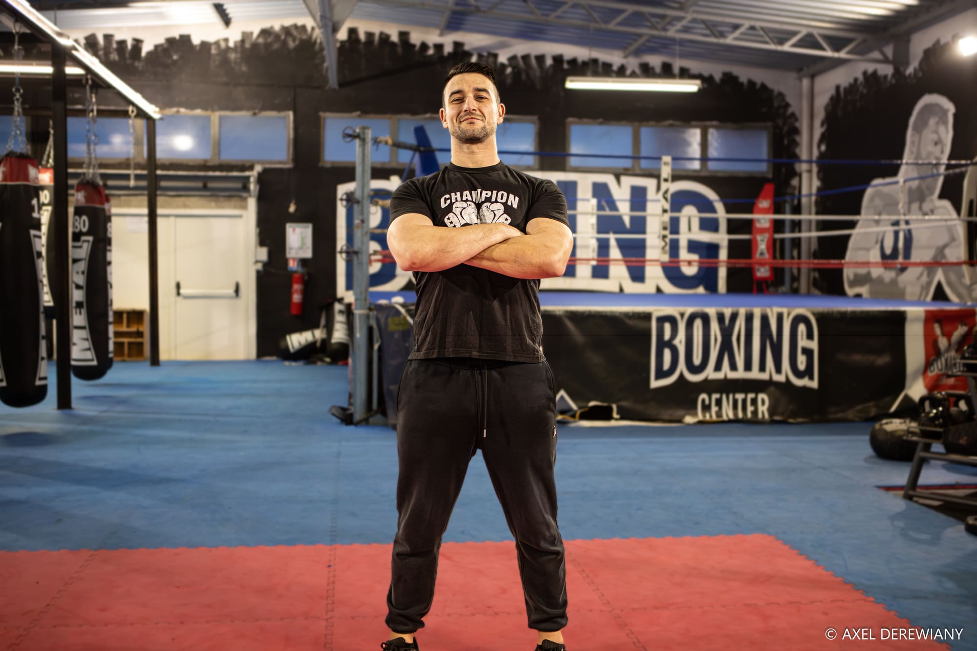 Un coach en t-shirt noir, bras croisés, debout sur le tapis rouge devant le ring et la banderole du club.