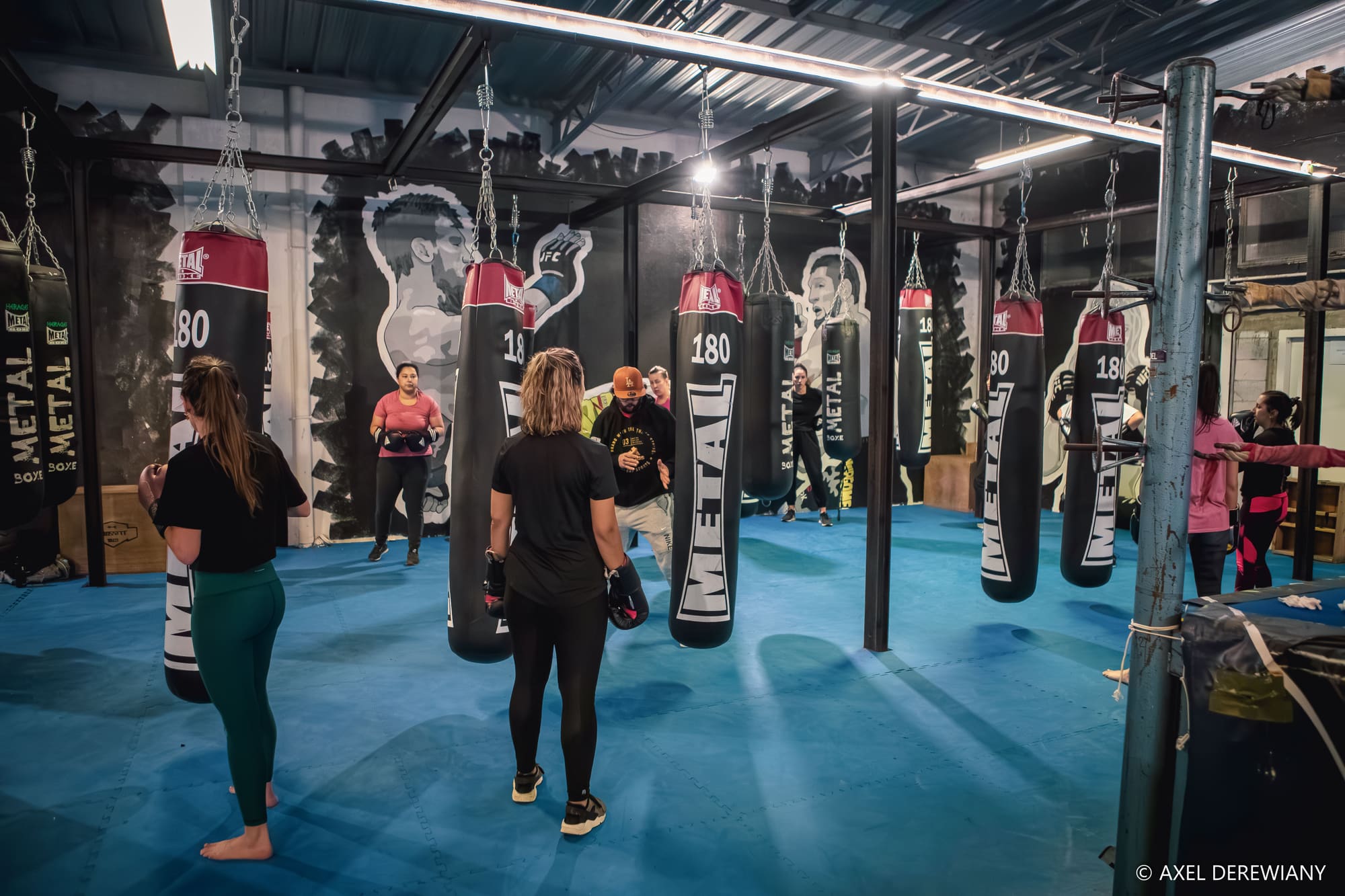 Un cours collectif au milieu des sacs Metal Boxe suspendus, sur le sol bleu, le coach au centre du groupe.
